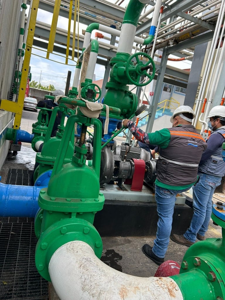 Workers inspecting large green industrial pump equipment with valves and pipes in a warehouse facility