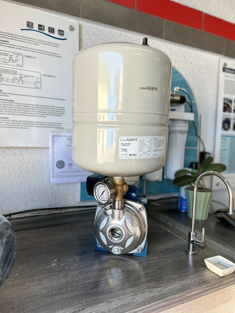Water pressure pump system with cream-colored storage tank mounted on metal base with pressure gauge, displayed on kitchen counter next to instruction manual